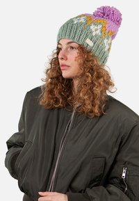 Knit beanie in muted green with yellow and white patterns, topped with a fluffy lavender pom-pom. Texture is thick and cozy.