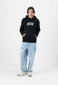 Sort hættetrøje med "HUF" logo, frontlomme, kombineret med lyseblå løstsiddende jeans og beige sneakers. Enkel hvid baggrund.