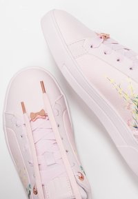 Baskets rose pâle en cuir texturé. Présentent des motifs floraux, des lacets en ruban et des accents en or rose avec un détail en forme de nœud.