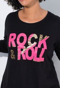Chemise à manches longues noire avec le mot "ROCK" en rose vif et le mot "ROLL" en imprimé léopard. Col rond et texture de tissu douce et lisse.