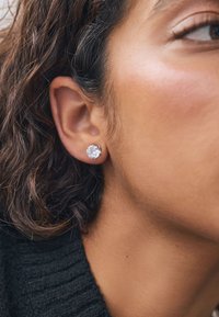 Silberne Ohrstecker mit klaren, runden Edelsteinen, vor gebräunter Haut und dunklem, strukturiertem Haar. Nahaufnahme, die die Ohrstecker hervorhebt.