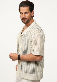 Strickshirt mit kurzen Ärmeln in Hellbeige und Creme mit einem geometrischen offenen Muster. Verfügt über einen spitzen Kragen und eine Brusttasche.