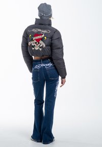 Black cropped puffer jacket with an embroidered skull design on the back. Blue flared jeans with white floral embroidery on the sides. Gray beanie.