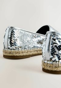 Chaussures espadrilles à sequins argentés avec semelles en jute tressé et semelles extérieures en caoutchouc, vues de l'arrière en angle sur un fond neutre.