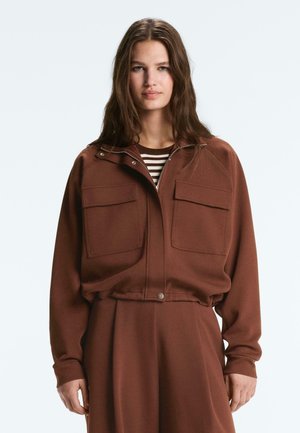 Mlada ženska z dolgimi lasmi nosi rjavo jakno z zadrgo in velikimi sprednjimi žepi ter ujemajoče se ohlapne hlače prek črtaste srajce.