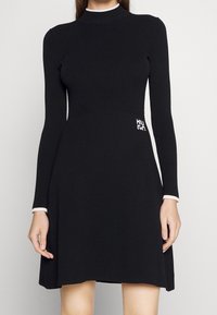Robe noire côtelée avec des manches longues, col montant, ourlet évasé et détail logo blanc sur le côté. Accents blancs contrastants aux poignets.