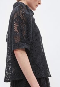 Μαύρο δαντελένιο μπλουζάκι με κοντά φουσκωτά μανίκια, που διαθέτει περίτεχνα λουλουδένια μοτίβα και φαρδιά εφαρμογή, αναδεικνύοντας ένα υφασμένο, δικτυωτό σχέδιο.