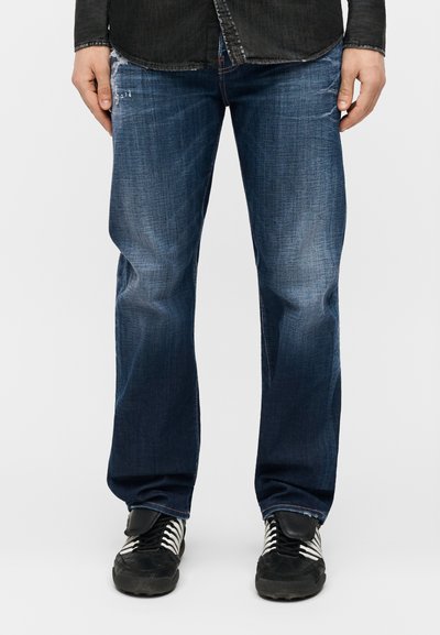 Homme portant un jean bleu foncé avec un léger effet délavé et une chemise noire, debout avec les mains détendues sur les côtés, baskets noires avec des lacets blancs.