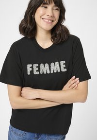 Tricou negru cu mâneci scurte, având cuvântul "FEMME" scris cu sclipici argintiu strălucitor. Gât rotund, croială lejeră și textură de material neted.