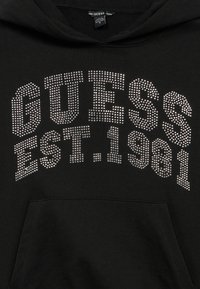 Hanorac negru cu textul „GUESS EST. 1981” în pietre strălucitoare argintii, cu buzunar tip cangur și textură moale a materialului.