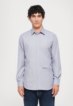 Young man with curly hair wearing a light blue button-up shirt with "DSQUARED2" logo and dark blue jeans, standing against a plain background.
