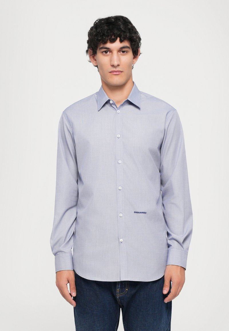 Junger Mann mit lockigem Haar, der ein hellblaues Hemd mit Knöpfen und dem "DSQUARED2"-Logo sowie dunkelblaue Jeans trägt, steht vor einem einfarbigen Hintergrund.