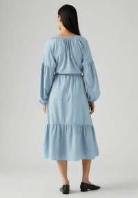 Vaaleansininen chambray-mekko, jossa on pitkät puffihihat, joustava vyötärö ja kerroksellinen hame. Lattialle ulottuva, rento istuvuus ja pyöreä pääntie.