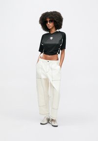 Μαύρο κοντό τοπ  με διαφανή υφή και λευκές λεπτομέρειες, σετ με χαλαρά λευκά παντελόνια με εμφανείς ραφές και τσέπες, ολοκληρωμένο με γκρι αθλητικά παπούτσια.