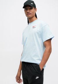 Gaiši zilas kokvilnas T-kreklu ar īsām piedurknēm un sarkanu Nike logo uz krūtīm, apvienots ar melniem sporta biksēm, kam ir balts Nike logo.
