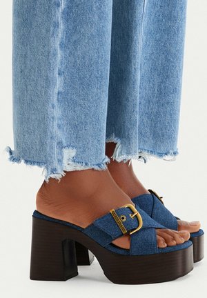 Pies con sandalias de plataforma de denim azul con hebillas doradas combinadas con vaqueros azul claro con dobladillo deshilachado.