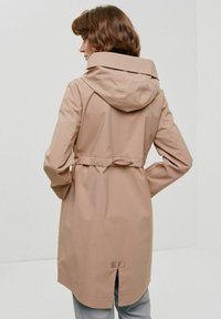Beżowa kurtka przeciwdeszczowa z dopasowanym kapturem, ściągniętą w talii i rozcięciem z tyłu. Gładki, wodoodporny materiał, długie rękawy.