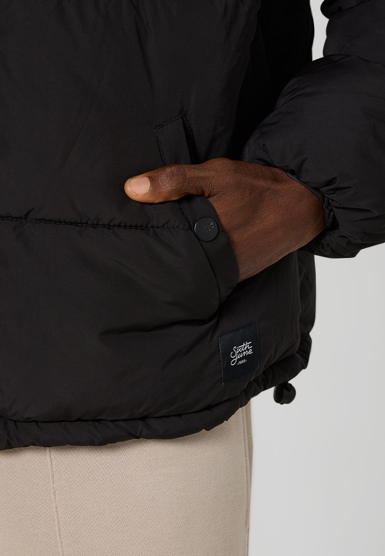 Czarna ocieplana kurtka z zamkiem błyskawicznym, elastycznymi mankietami i naszywką z logo. Materiał wydaje się gładki i wodoodporny.