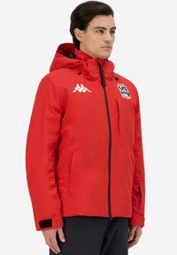 Giacca impermeabile rossa con cappuccio, zip e polsini in velcro. Presenta un logo sul petto e una superficie liscia.
