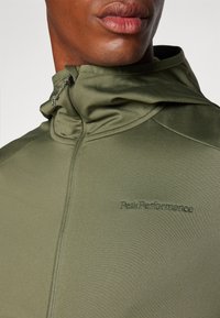Olive green athletic jacket with a high collar, features a zip closure and logo embroidered on the chest, smooth texture and fitted design.