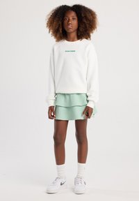 Sudadera blanca con texto verde, combinada con una falda corta en tonos verde claro. Lleva puesta unas zapatillas blancas y calcetines blancos por encima del tobillo.