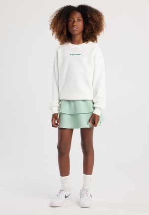 Sudadera blanca con texto verde, combinada con una falda corta en tonos verde claro. Lleva puesta unas zapatillas blancas y calcetines blancos por encima del tobillo.