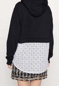Sudadera negra con capucha y un panel inferior blanco perforado que presenta un patrón triangular, combinada con una falda negra con detalles de cadenas.
