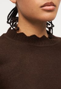 Κοντινό πλάνο ενός ατόμου που φοράει σκούρο καφέ πλεκτό πουλόβερ με διακριτικό κυματιστό γιακά και πλεξούδες που ακουμπούν στον λαιμό.