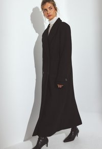 Dlhý čierny vlnený kabát s dvojradovým zapínaním, širokými klopami a bočnými vreckami. Nosí sa cez biele rolákové tričko a je skombinovaný s čiernymi členkovými čižmami.