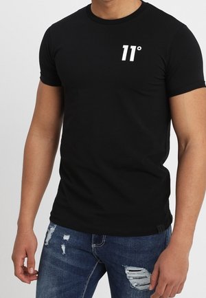 Osoba ubrana w czarną dopasowaną koszulkę z nadrukiem "11°" na piersi oraz podniszczone niebieskie dżinsy.