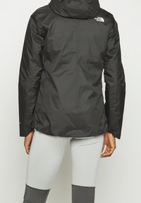 Černá nepromokavá bunda s kapucí. Má hladkou texturu a logo na horní části zad. Nosen�á s světle šedými legínami.