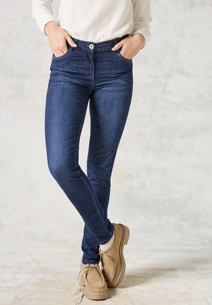 Jeans skinny in denim blu scuro con chiusura frontale a bottoni e zip, cinque tasche e dettagli di cucitura minimi. Abbinati a scarpe marroni chiare.