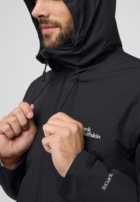 Zwarte waterdichte jas met een goed passende capuchon, verstelbare trekkoorden en klittenbandsomen; voorzien van een logo op de borst.