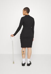 Svart långärmad topp med volangkrage, ihop med en svart åtsittande kjol. Visar vita strumpor och kraftiga svarta skor. Tydlig käpp i närheten.