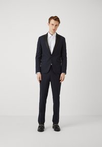 Tuxedo blu navy con rever a scialle, vestibilità slim e pantaloni abbinati. Accompagnato da una camicia bianca e scarpe nere con dettagli in oro.