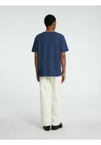 Mörkblå t-shirt med korta ärmar och rundad halsringning, matchad med vita byxor och svarta skor, sedd från baksidan.