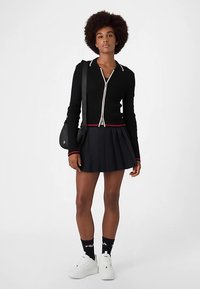 Musta ribbineuloksinen neuletakki hopeanvärisellä vetoketjulla ja punaisella reunuksella, yhdistettynä laskostettuun mustaan hameeseen, valkoisiin lenkkareihin ja mustiin logo-sukkiin.