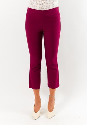 Donna con pantaloni corti magenta, tacchi alti bianchi appuntiti e una maglia bianca in pizzo, in piedi su uno sfondo bianco semplice.