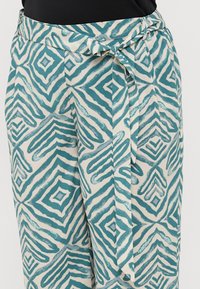 Patterned loose-fit trousers in cream with teal geometric print, featuring a tied waist and smooth fabric texture.