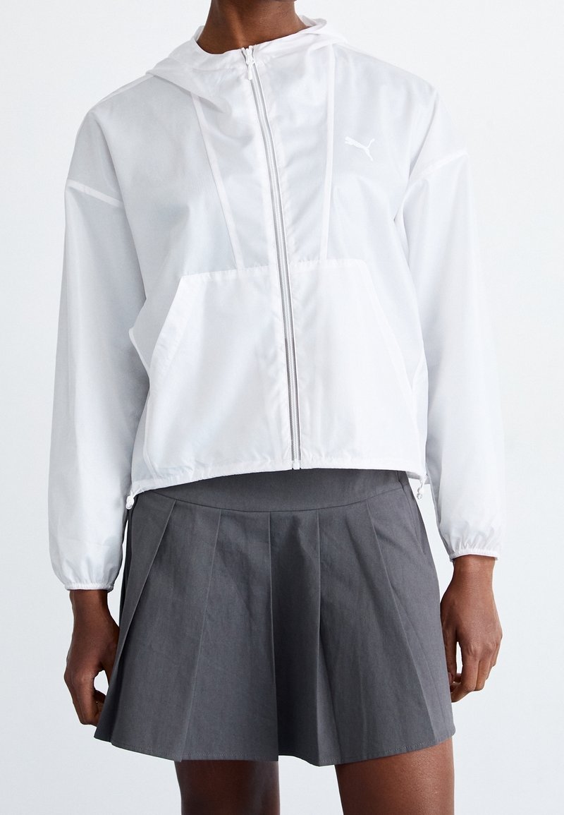 Biała kurtka z kapturem wykonana z lekkiego materiału, wyposażona w przedni zamek, dużą kieszeń i subtelne logo. Połączona z szarą plisowaną spódnicą.