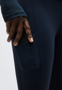 Χέρι με σκούρο χρώμα δέρματος που βάζει ένα μαύρο smartphone στην πλαϊνή τσέπη σκούρου μπλε εφαρμοστών αθλητικών κολάν.