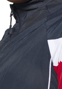 Kevyt, tumman laivaston sininen tuulenpitävä takki, jossa on korkea kaulus, puoli-vetoketjukiinnitys ja kontrastiväriset punaiset ja valkoiset paneelit hihoissa.