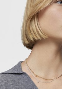 Collane a strati con una catena d'oro e un piccolo ciondolo, insieme a una catena d'argento con un accento luccicante. Indossate su un capo in maglia grigia.