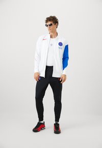 Hvid zip-up jakke med blå ærmer og røde detaljer, sort tætsiddende bukser og rød-sorte sneakers. Solbriller fuldender det sporty look.