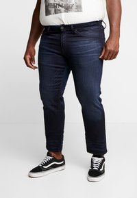 Dark denim jeans with a slim fit, featuring five pockets and a button closure. Paired with black and white patterned sneakers.