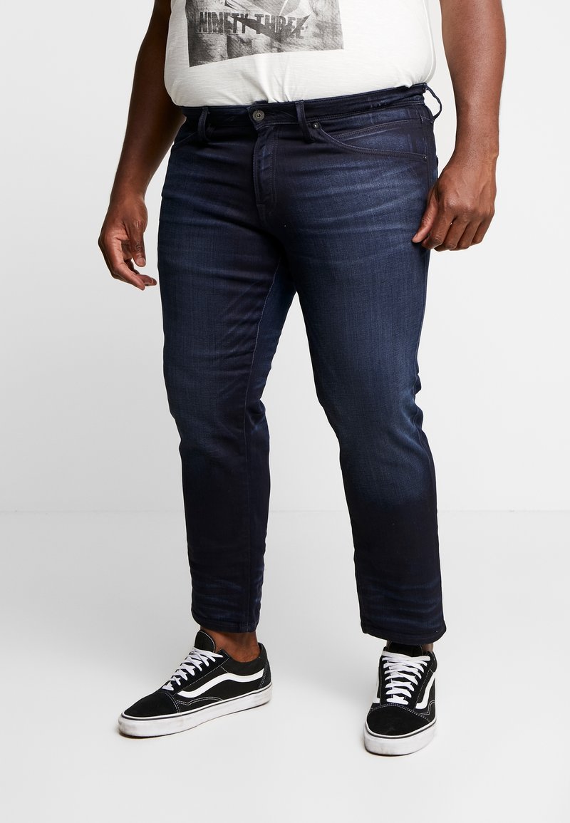 Dark denim jeans with a slim fit, featuring five pockets and a button closure. Paired with black and white patterned sneakers.