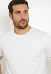 Camiseta de algodón blanca con un escote redondo, mangas cortas y un logo sutil de marca en beige claro en el pecho. Textura suave.