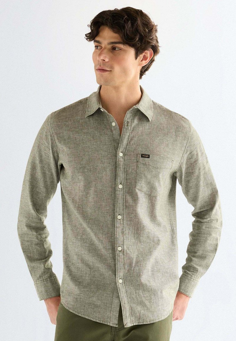 Junger Mann mit dunklen Haaren, der ein olivgrünes Hemd mit langen Ärmeln und Brusttasche trägt und mit den Händen in den Taschen steht.