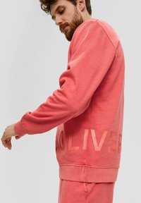 Sweatshirt corail avec un col rond, des poignets et un ourlet côtelés. Présente un texte « OLIVER » audacieux avec une finition brillante sur le côté gauche. Texture douce.