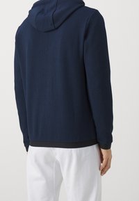 Felpa con cappuccio di un blu navy scuro in tessuto texturizzato, con orlo e polsini a coste neri. Dettaglio di cucitura visibile sul retro.